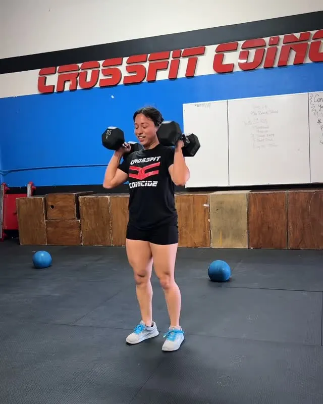 A woman in a Coincide shirt training with two dumbbells at her shoulders