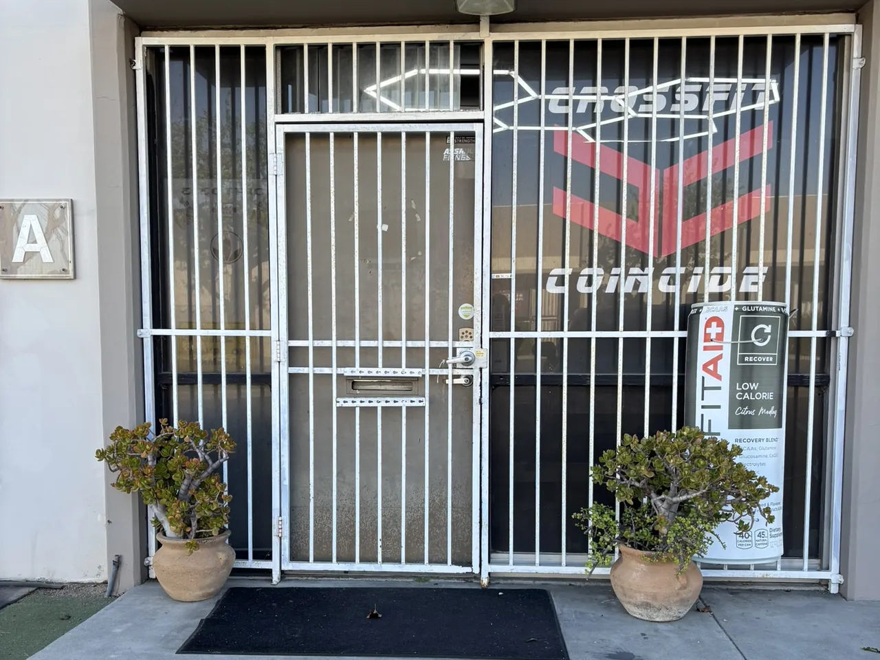 Our door: a white metal gate with the red CrossFit Coincide logo on the glass behind it and an “A” on the wall beside it